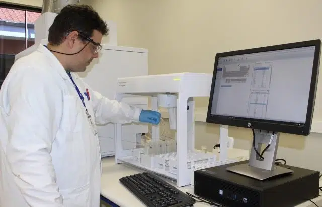 Um cientista de jaleco branco e óculos de proteção opera uma máquina de análise química, enquanto monitora os dados da amostra de solo em um computador ao lado.