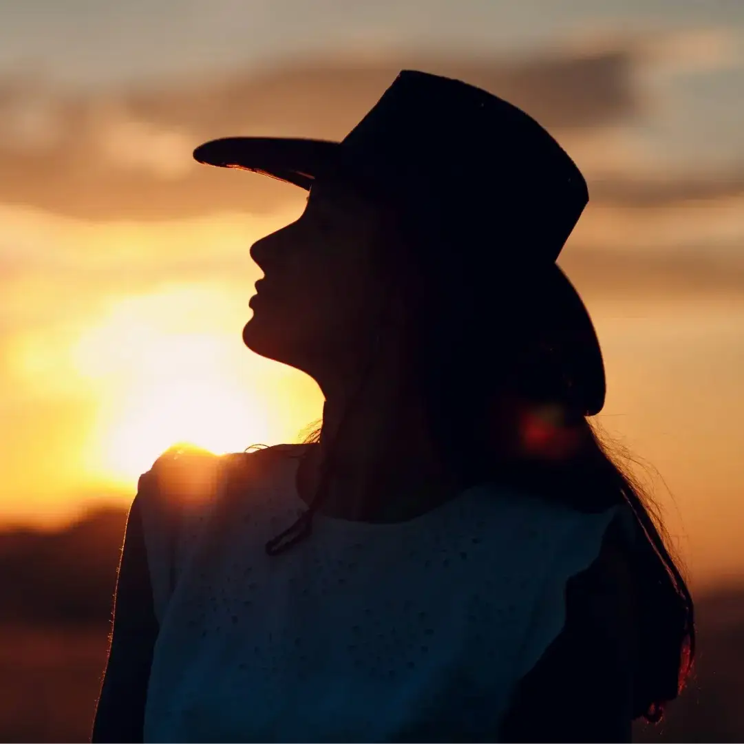 Perfil de uma mulher em silhueta, olhando para o horizonte durante o entardecer. O sol poente cria uma aura dourada ao redor de seu chapéu e vestimenta, transmitindo uma atmosfera de reflexão e conexão com a terra.