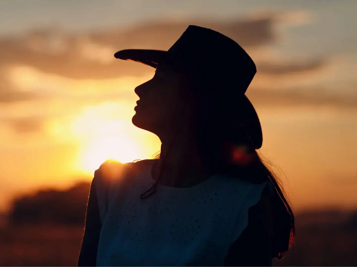 Perfil de uma mulher em silhueta, olhando para o horizonte durante o entardecer. O sol poente cria uma aura dourada ao redor de seu chapéu e vestimenta, transmitindo uma atmosfera de reflexão e conexão com a terra.