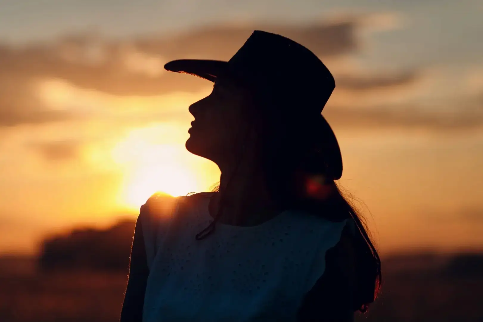 Perfil de uma mulher em silhueta, olhando para o horizonte durante o entardecer. O sol poente cria uma aura dourada ao redor de seu chapéu e vestimenta, transmitindo uma atmosfera de reflexão e conexão com a terra.